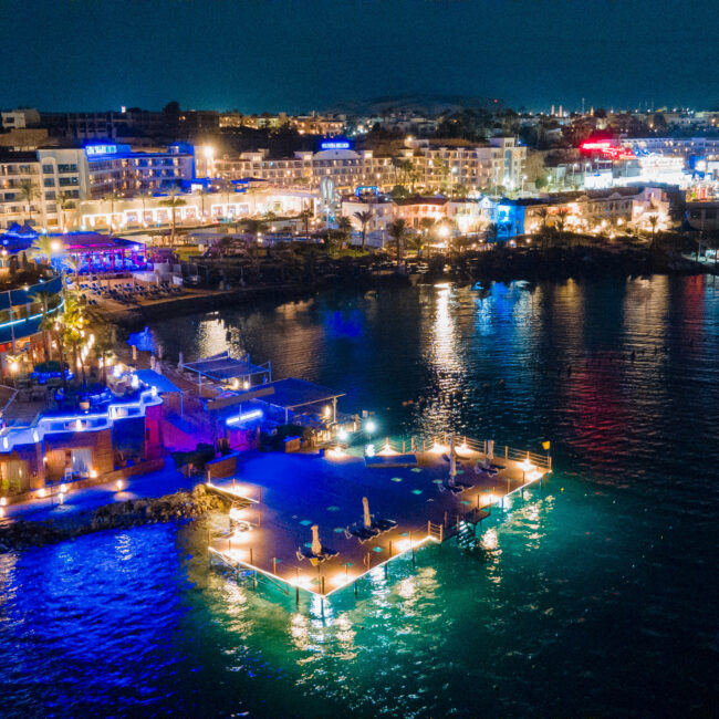 Night aerial view of a brightly lit harborfront with hotels, restaurants, and boats along a calm bay at dusk.
