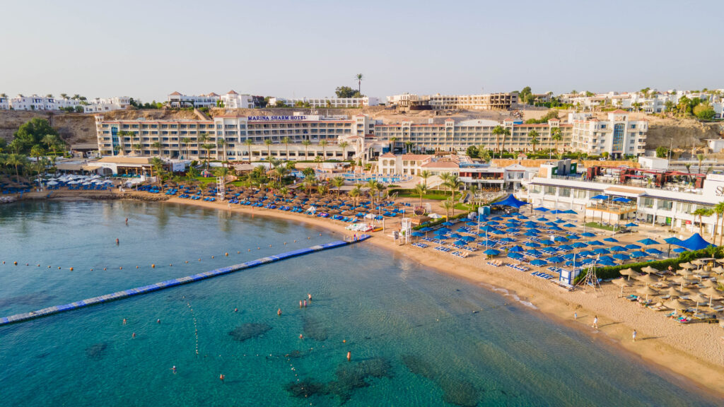 Aerial view of Marina Sharm Hotel complex along a sandy beach with rows of blue sun loungers and umbrellas by turquoise water.