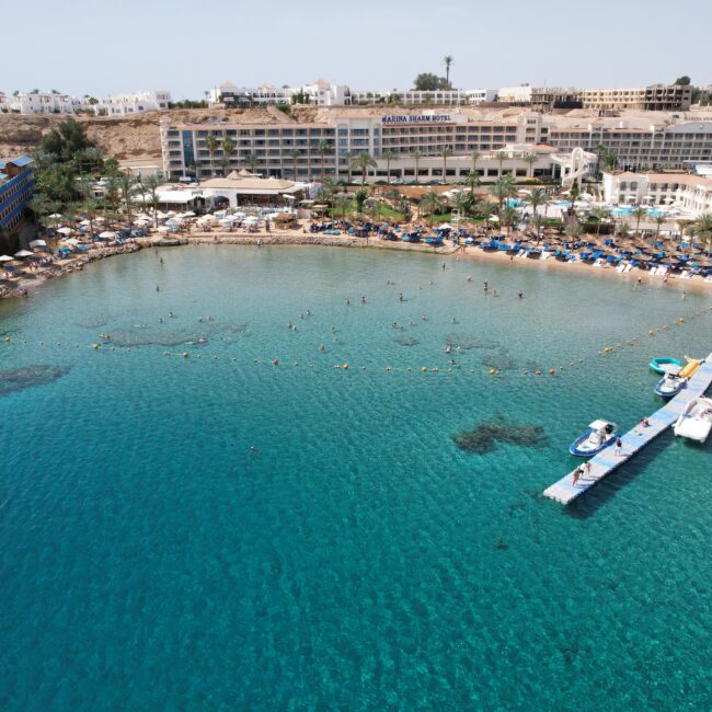 Aerial view of a seaside resort with white buildings, palm trees, a sandy beach, and turquoise water with sun loungers along the shore.